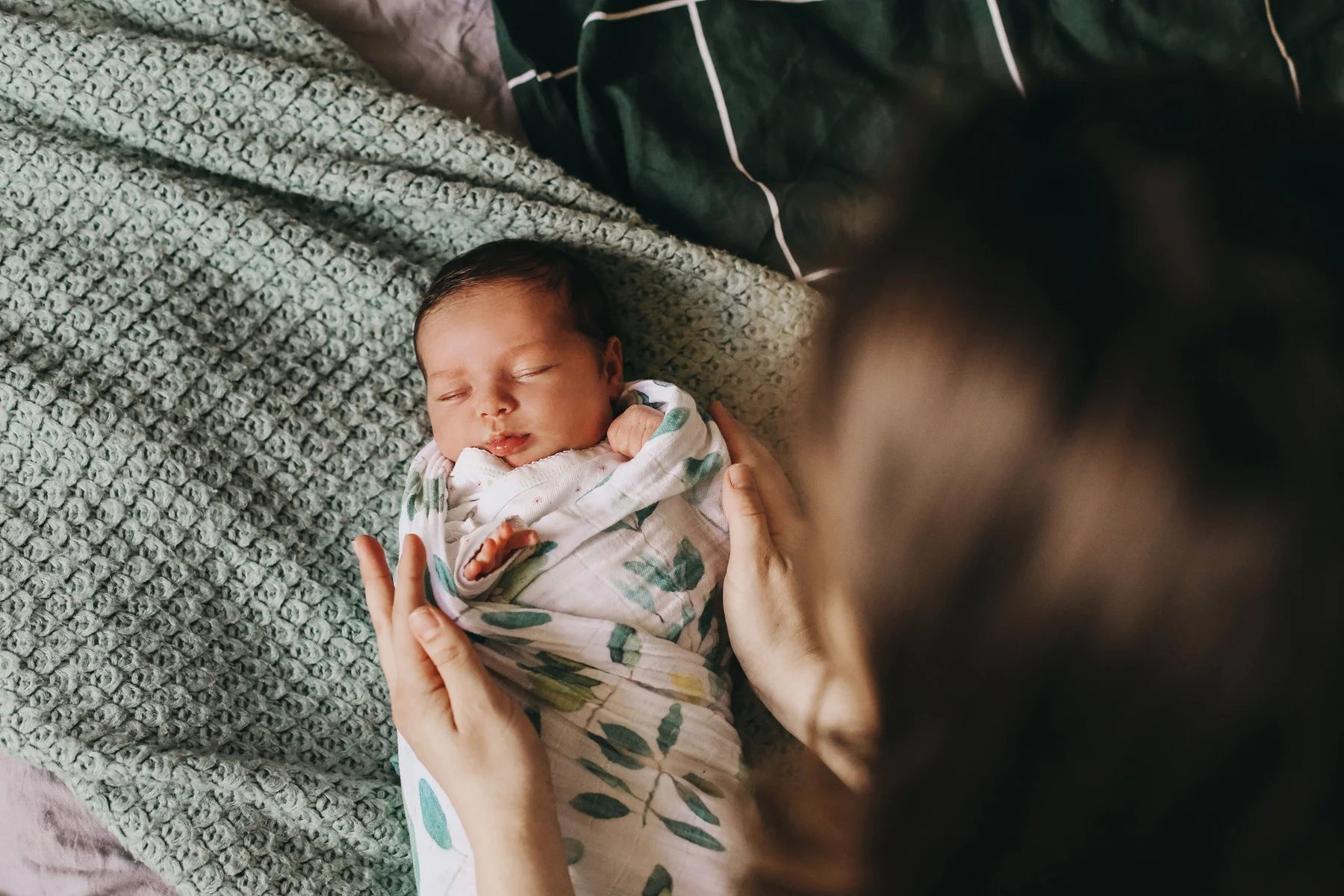 A newborn swaddled in a leaf-print wrap, held by a parent