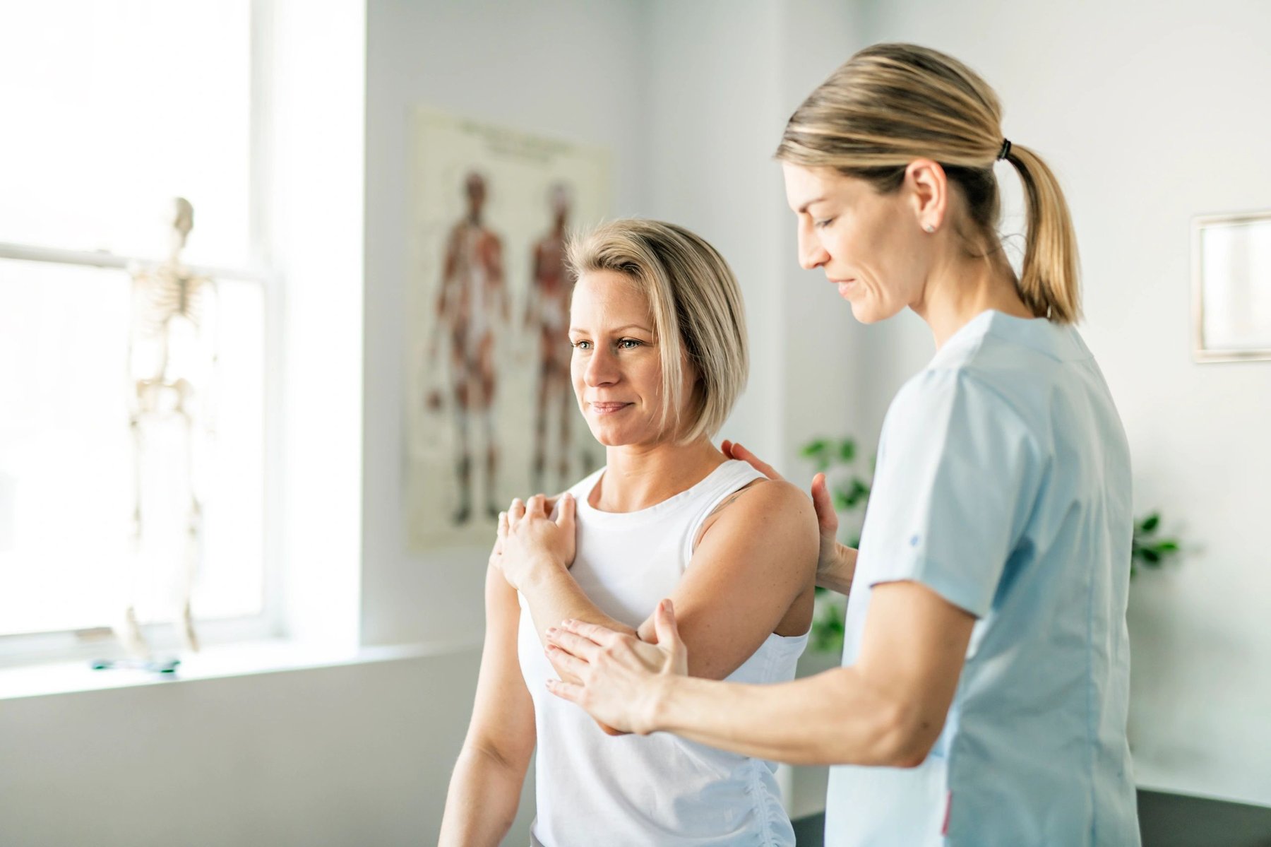 Physiotherapist guiding shoulder range of motion in a clinic with an anatomy poster
